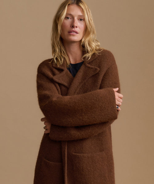 A woman wearing a long, oversized brown wool coat with wide lapels and a tie belt, posed against a solid beige studio background.
