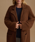A woman wearing a plush, oversized brown coat over a dark navy top, photographed against a solid warm beige background.
