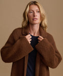 A woman wearing a brown fuzzy textured coat over a black top, posing against a solid beige background.