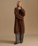 A woman stands against a plain beige studio background wearing a long brown fuzzy coat over a dark top and striped pants.