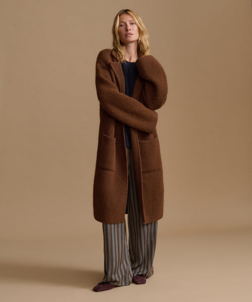 A woman stands against a plain beige studio background wearing a long brown fuzzy coat over a dark top and striped pants.