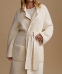 A woman wearing a cream-colored, fuzzy belted coat over a matching cream top, set against a solid beige background.