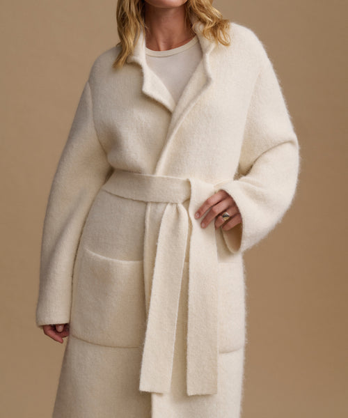 A woman wearing a cream-colored, fuzzy belted coat over a matching cream top, set against a solid beige background.