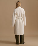 A woman viewed from the back wearing a long cream-colored belted coat over dark olive green trousers, set against a solid warm beige studio background.