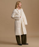 A woman modeling a long cream-colored bouclé coat with a matching tie belt, worn over a light top and dark olive green wide-leg trousers, set against a solid beige studio background.