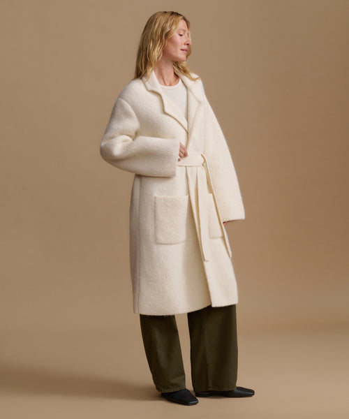 A woman modeling a long cream-colored bouclé coat with a matching tie belt, worn over a light top and dark olive green wide-leg trousers, set against a solid beige studio background.