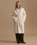 A woman modeling a cream-colored, oversized belted coat over a white top and dark olive green trousers, standing against a solid beige studio background.