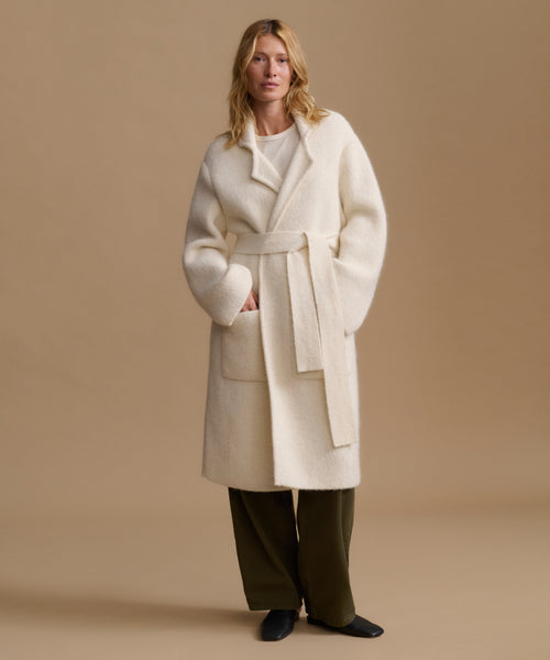 A woman modeling a cream-colored, oversized belted coat over a white top and dark olive green trousers, standing against a solid beige studio background.