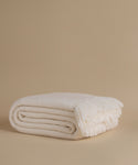 A folded cream-colored knit throw blanket with fringed ends, displayed on a solid beige background.