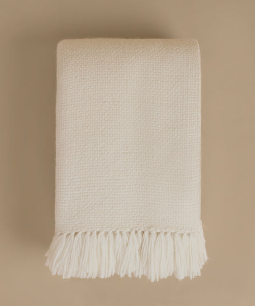 A folded cream-colored woven throw blanket with fringed edges, displayed against a solid beige background.