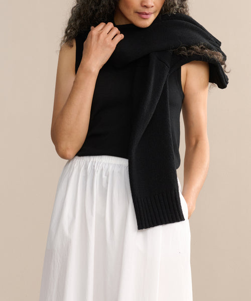 Wearing a sleeveless black top and the Lauren Skirt, a person stands against a beige background with a black sweater draped over their shoulders.