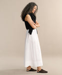 A woman with curly hair stands in profile against a plain background, wearing a black sleeveless top, the Lauren Skirt as her wardrobe staple, a black sweater draped over her shoulders, and black woven sandals.