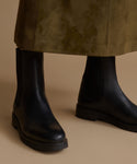 A close-up of a person wearing black Chelsea boots with a dark olive green skirt against a warm beige background.