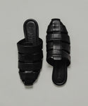 The Leather Camilla Mule is a pair of black leather slip-on sandals with wide interwoven straps and flat soles, displayed on a plain light background.