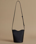 A black leather bucket bag with a long shoulder strap, photographed against a solid beige background.