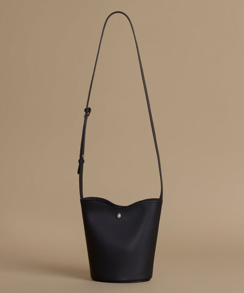 A black leather bucket bag with a long shoulder strap and a small silver clasp, set against a solid beige background.