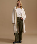 A woman stands against a warm beige studio background wearing a long cream fuzzy coat over a cream top, olive green wide-leg pants, and black shoes.