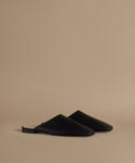A pair of black leather mule flats with a pointed toe, displayed on a warm beige studio background.