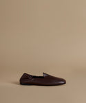 A single dark brown leather slip-on loafer shoe shown in profile against a solid beige studio background.