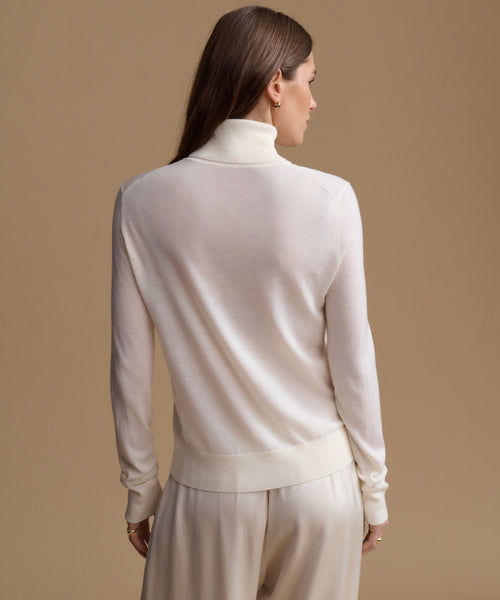 A woman shown from the back wearing a cream-colored knit mock neck sweater and matching knit trousers against a solid beige studio background.