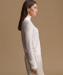 A woman in side profile wearing a cream-colored mock neck knit sweater and matching skirt against a solid beige background.