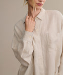 A person wears the loose, beige Linen Colby Shirt with a chest pocket, holding the collar with one hand. Their face is partly visible and hair tied back—ideal for relaxed summer style.