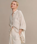 A woman with blonde hair in a low bun models the oversized Linen Colby Shirt in light beige with rolled sleeves, paired with cream pants. She stands against a neutral backdrop, one hand in her pocket—a perfect summer style.