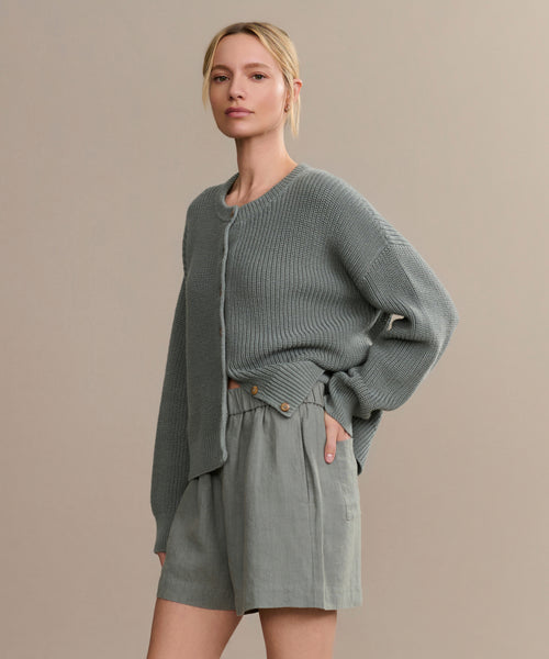 A woman with blonde hair in a low ponytail wears the Linen Logan Short and a loose, textured gray cardigan, creating an effortless warm-weather look against a plain beige background.