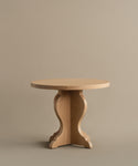 The Lombard Side Table features a round, solid white oak design with a smooth top and decorative pedestal base, blending vintage-inspired elegance and timeless craftsmanship against a plain beige background.