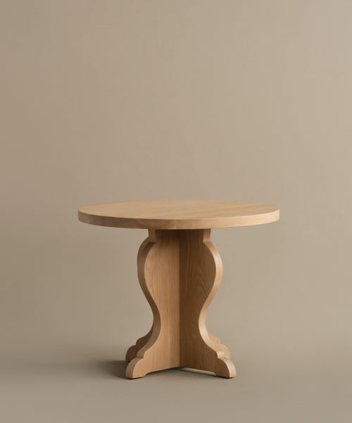 The Lombard Side Table features a round, solid white oak design with a smooth top and decorative pedestal base, blending vintage-inspired elegance and timeless craftsmanship against a plain beige background.
