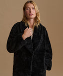A woman wearing a black shearling coat over a light beige top, standing against a solid beige background.