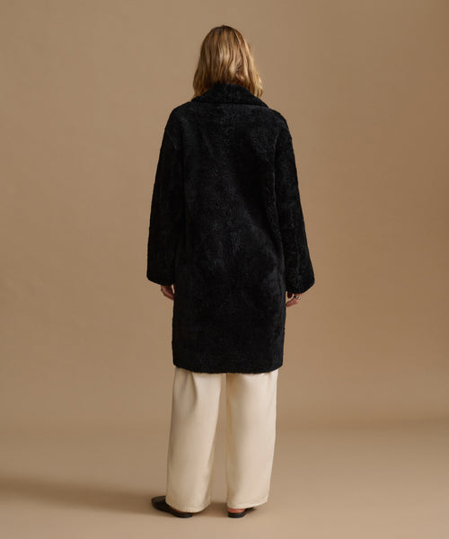 A model viewed from the back wearing a black shearling coat over cream wide-leg trousers, set against a solid warm beige studio background.