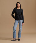A woman modeling a black long-sleeve crewneck top with light-wash blue jeans and black flats, standing against a solid beige studio background.