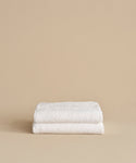 Two neatly folded Loom Bath Sheets, crafted from a luxurious cotton modal blend, are stacked on top of each other against a plain beige background.