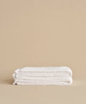 The Loom Bath Sheet, ultra-soft and white, is neatly folded on a neutral beige surface with a plain beige background.