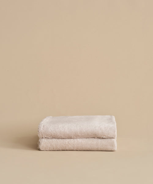 Two neatly folded, super-soft Loom Bath Sheets in beige are stacked atop each other against a plain beige background.