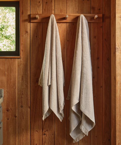Two Loom Bath Sheets in light beige hang side by side on wooden pegs against a rustic wall, softly illuminated by natural light from a nearby window.