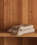 Two Loom Bath Sheets in soft beige cotton-modal are neatly folded and stacked on a wooden shelf, set against vertical wooden planks.