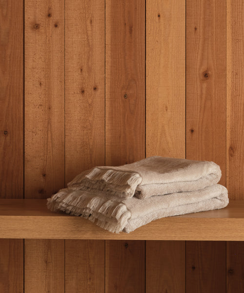 Two Loom Bath Sheets in soft beige cotton-modal are neatly folded and stacked on a wooden shelf, set against vertical wooden planks.