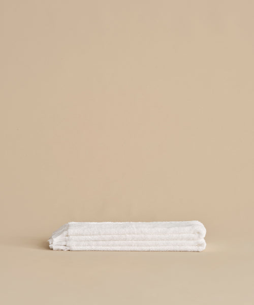 The Loom Hand Towel, ultra-soft and neatly folded in classic white, is displayed on a smooth beige surface with a coordinating beige background—an essential for any bathroom.