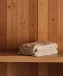 A stack of Loom Hand Towels in beige cotton-modal with fringed edges sits on a wooden shelf against vertical wooden panels, making them an essential for any bathroom.