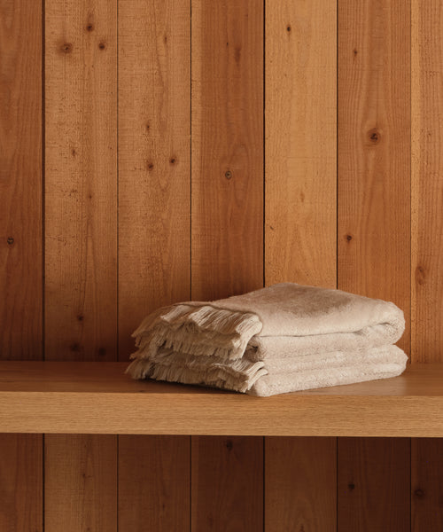 A stack of Loom Hand Towels in beige cotton-modal with fringed edges sits on a wooden shelf against vertical wooden panels, making them an essential for any bathroom.