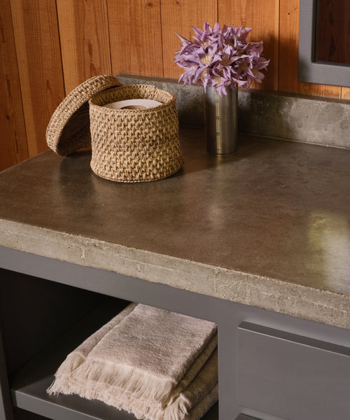 On a concrete countertop sits a lidded woven basket with toilet paper and a metal vase of purple flowers; below, Loom Hand Towels—everyday bathroom essentials—are neatly folded on a gray shelf against a wood-paneled wall.