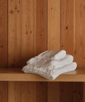 Three neatly folded Loom Wash Cloths, made from soft cotton modal, are stacked on a wooden shelf with vertical wooden planks in the background—an essential touch for any bathroom.