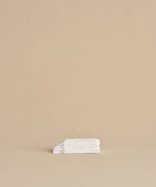 The Loom Wash Cloth, made from white cotton modal, is neatly folded on a beige surface with a plain beige background, showcasing a minimalist and clean bathroom essential.