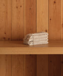 A neatly folded stack of super-soft beige Loom Wash Cloths sits on a wooden shelf with vertical wooden panels in the background—a true bathroom essential.
