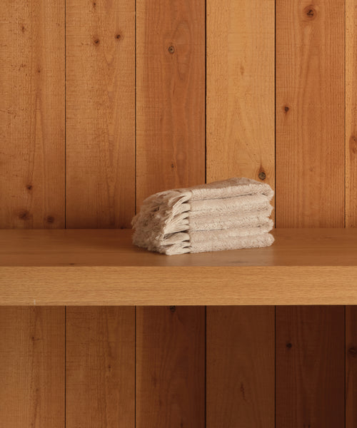 A neatly folded stack of super-soft beige Loom Wash Cloths sits on a wooden shelf with vertical wooden panels in the background—a true bathroom essential.