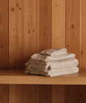 A neatly folded stack of super-soft Loom Wash Cloths in beige with fringed edges sits on a wooden shelf against vertical wood panels—a true bathroom essential.