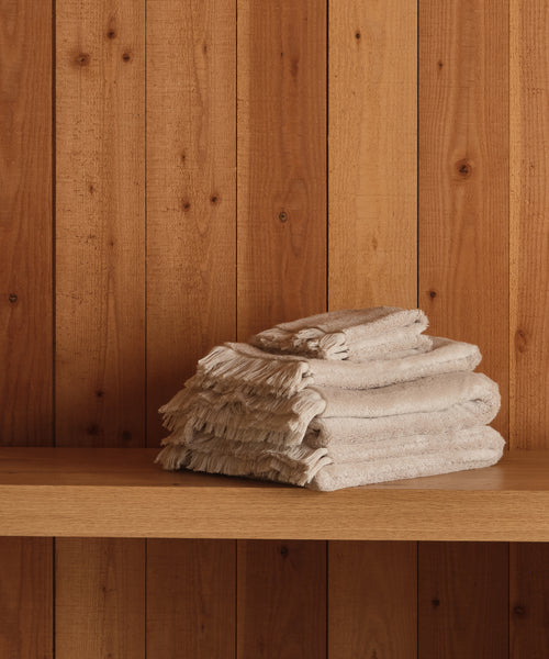 A neatly folded stack of super-soft Loom Wash Cloths in beige with fringed edges sits on a wooden shelf against vertical wood panels—a true bathroom essential.