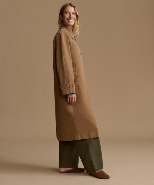 A woman modeling a long tan coat over olive green wide-leg pants and brown suede shoes, standing against a solid tan studio background.